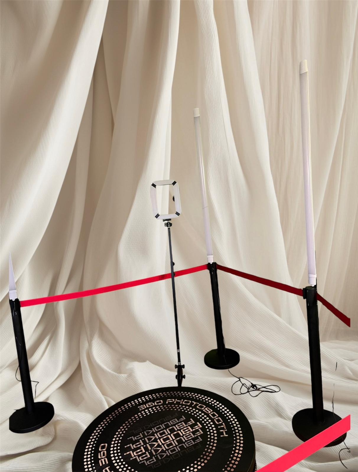 Okrągła platforma 360 z napisem '360 PHOTOBOOTH' i logo 'FBUDKI.PL', otoczona czarnymi słupkami z białymi lampami i czerwoną taśmą na tle białej, drapowanej tkaniny.