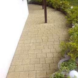 Żółta ścieżka ułożona z kostki brukowej prowadząca wzdłuż białej ściany budynku i obrzeża z roślinnością.