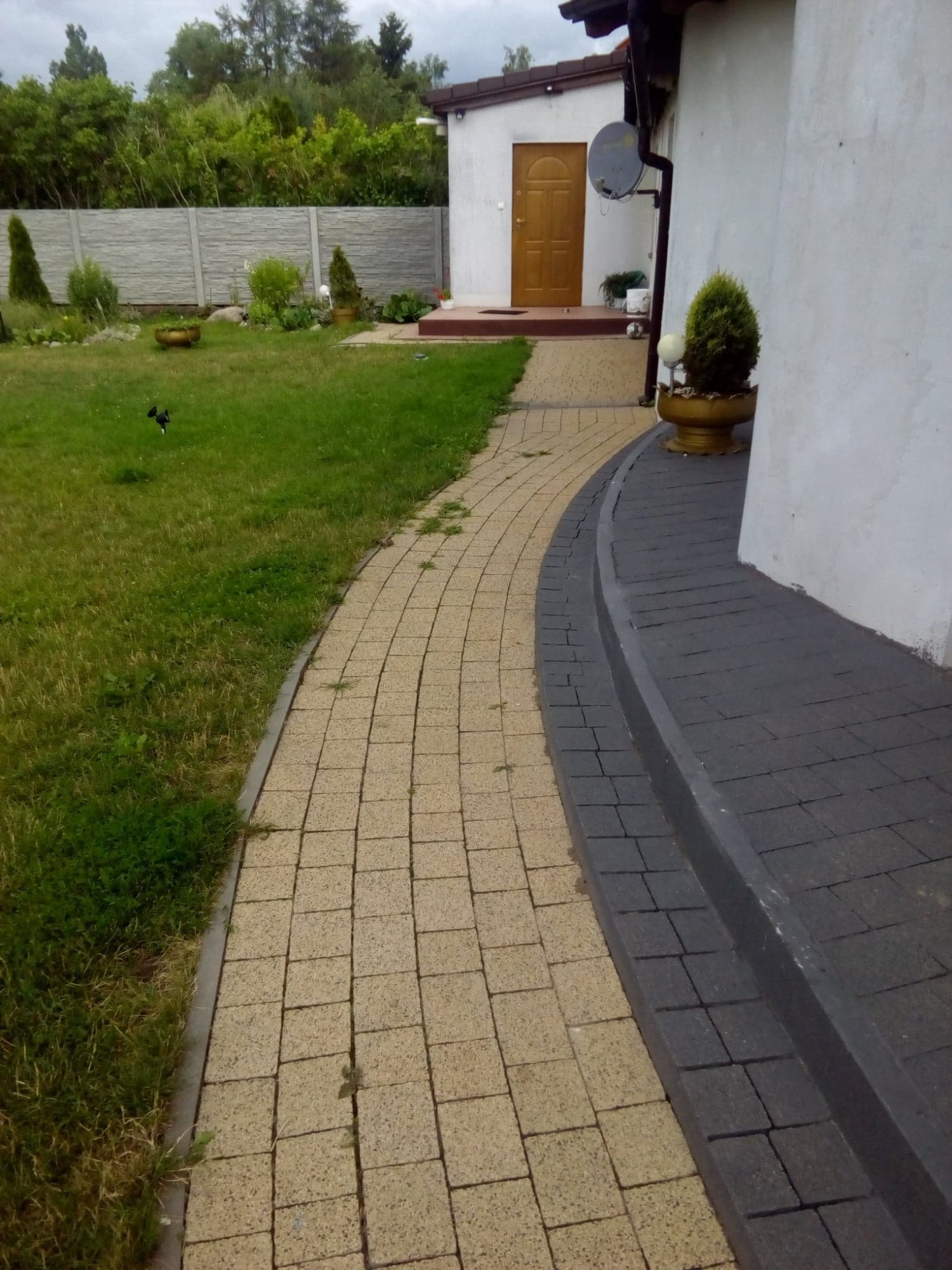 Zakrzywiony chodnik z beżowej kostki brukowej przechodzący w ciemnoszarą kostkę przy białym budynku, widok na zadbany trawnik i ogrodzenie w tle.