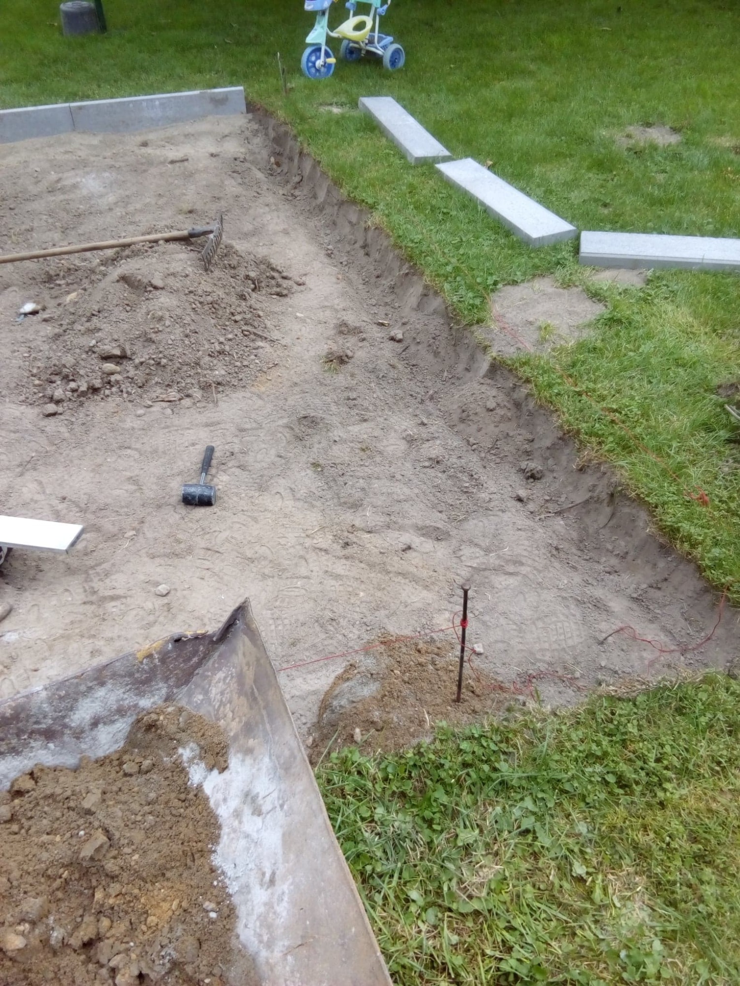 Przygotowanie terenu pod ścieżkę: wykopana ziemia, wyrównany teren, częściowo ułożone obrzeża, widoczne narzędzia - grabie i młotek brukarski, taczka z ziemią w rogu kadru, w tle dziecięcy...