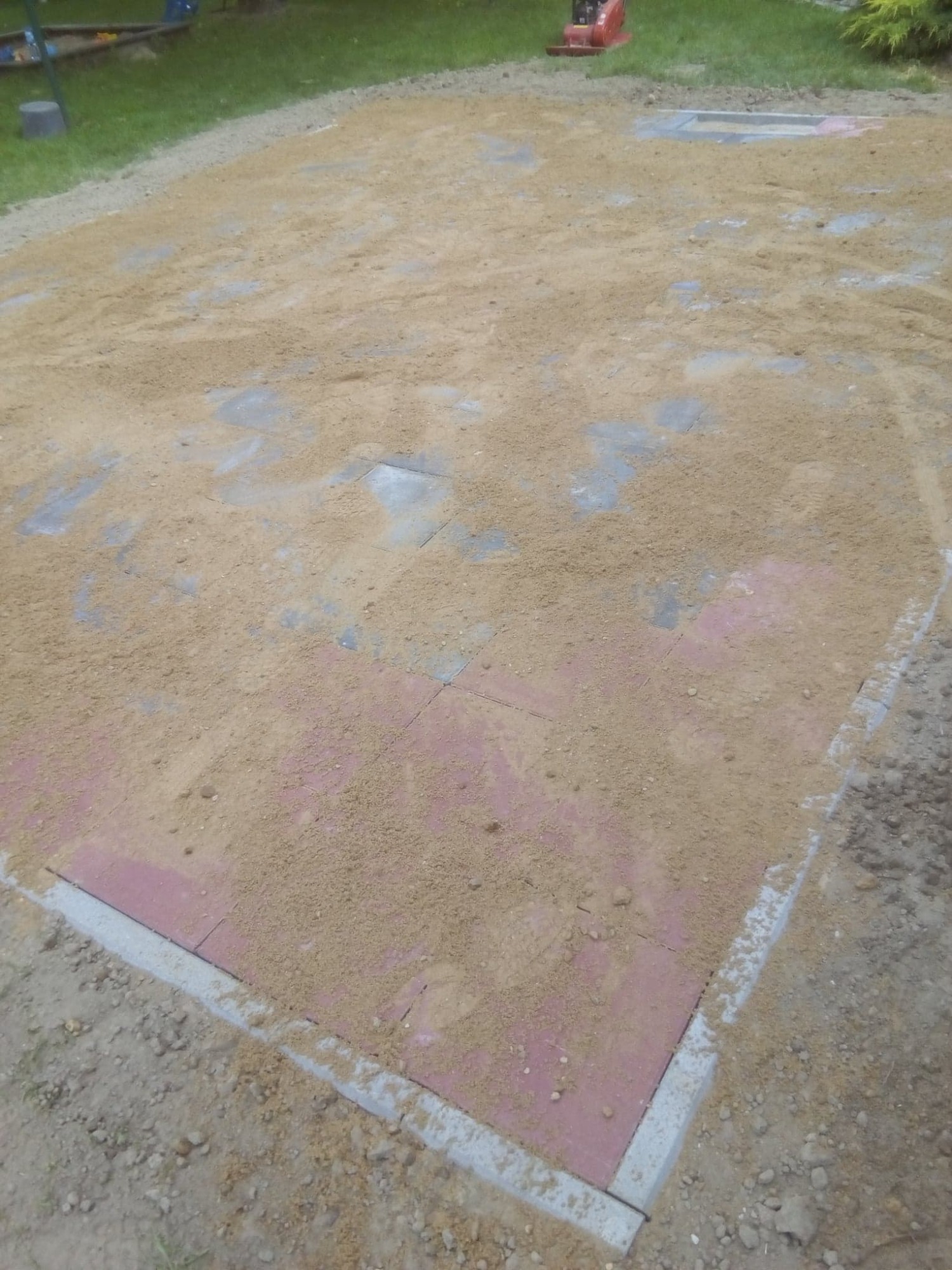 Przygotowany teren pod ułożenie nawierzchni, częściowo pokryty piaskiem, z widocznymi fragmentami kostki brukowej i obrzeżami. W tle zagęszczarka.