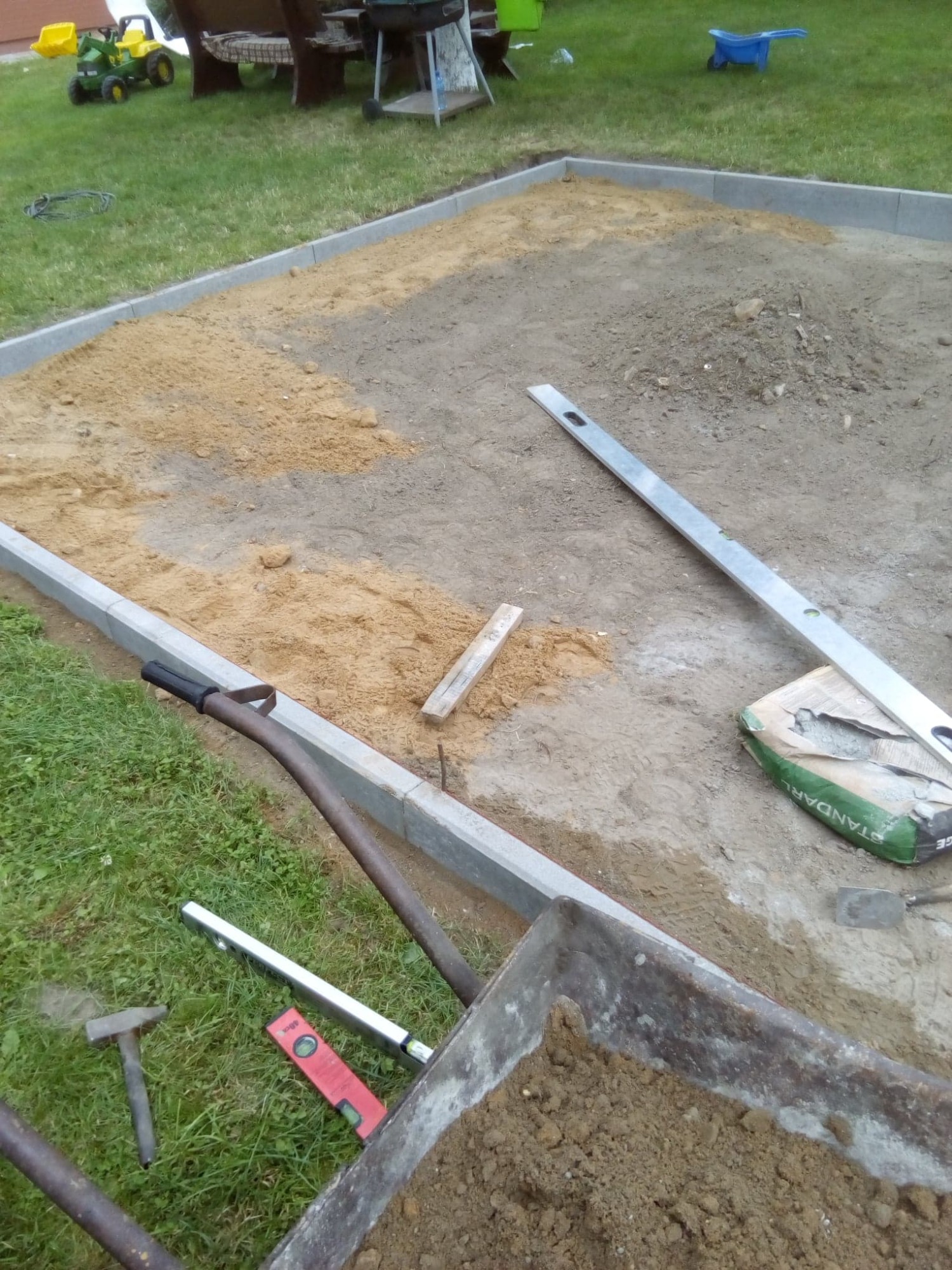 Przygotowanie podłoża pod nawierzchnię: obrzeża z kostki betonowej wyznaczają obszar, piasek częściowo rozsypany, widoczne narzędzia i materiały budowlane, w tle zabawki ogrodowe.