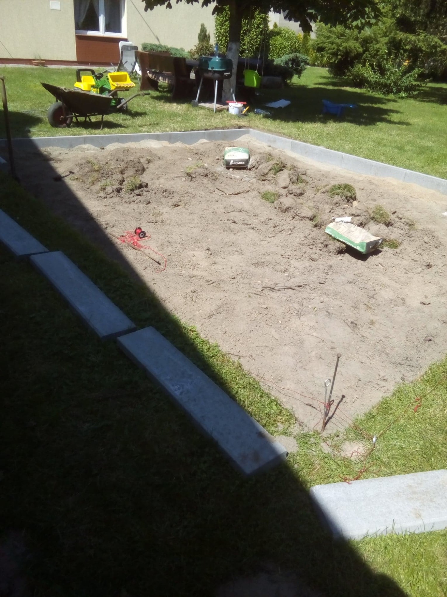 Przygotowanie podłoża pod kostkę brukową: wyrównany teren otoczony obrzeżami, widoczne szpilki geodezyjne i sznurek pomiarowy, w tle taczka z zabawkami.