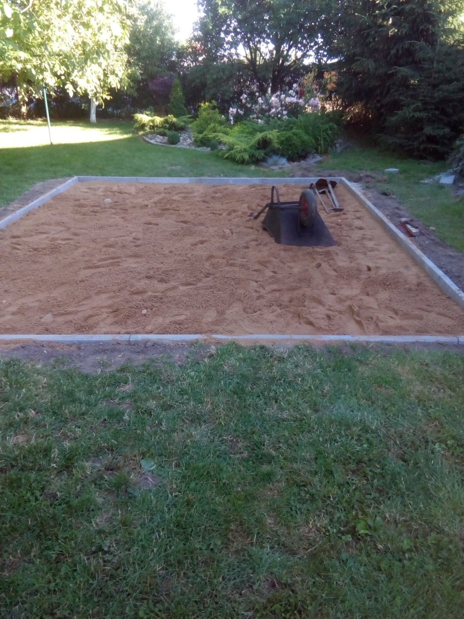 Przygotowany teren pod budowę z obramowaniem z szarej kostki, wypełniony piaskiem, z widoczną taczką i łopatą. W tle trawnik i ozdobne krzewy.