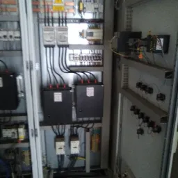 Otwarta szafa elektryczna z widocznymi bezpiecznikami, przewodami, transformatorami i oznaczeniem ostrzegawczym wysokiego napięcia.