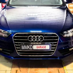 Przód granatowego samochodu Audi z widoczną tablicą rejestracyjną 'MMKD GARAGE' na tle warsztatu samochodowego.