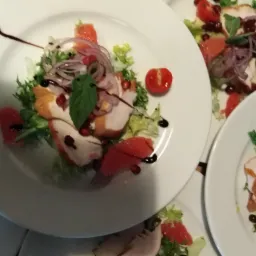 Elegancko ułożona sałatka z grillowanym kurczakiem, grejpfrutem, granatem, czerwoną cebulą i bazylią, polana sosem balsamicznym, na białym talerzu. Kilka talerzy z podobną kompozycją w tle.