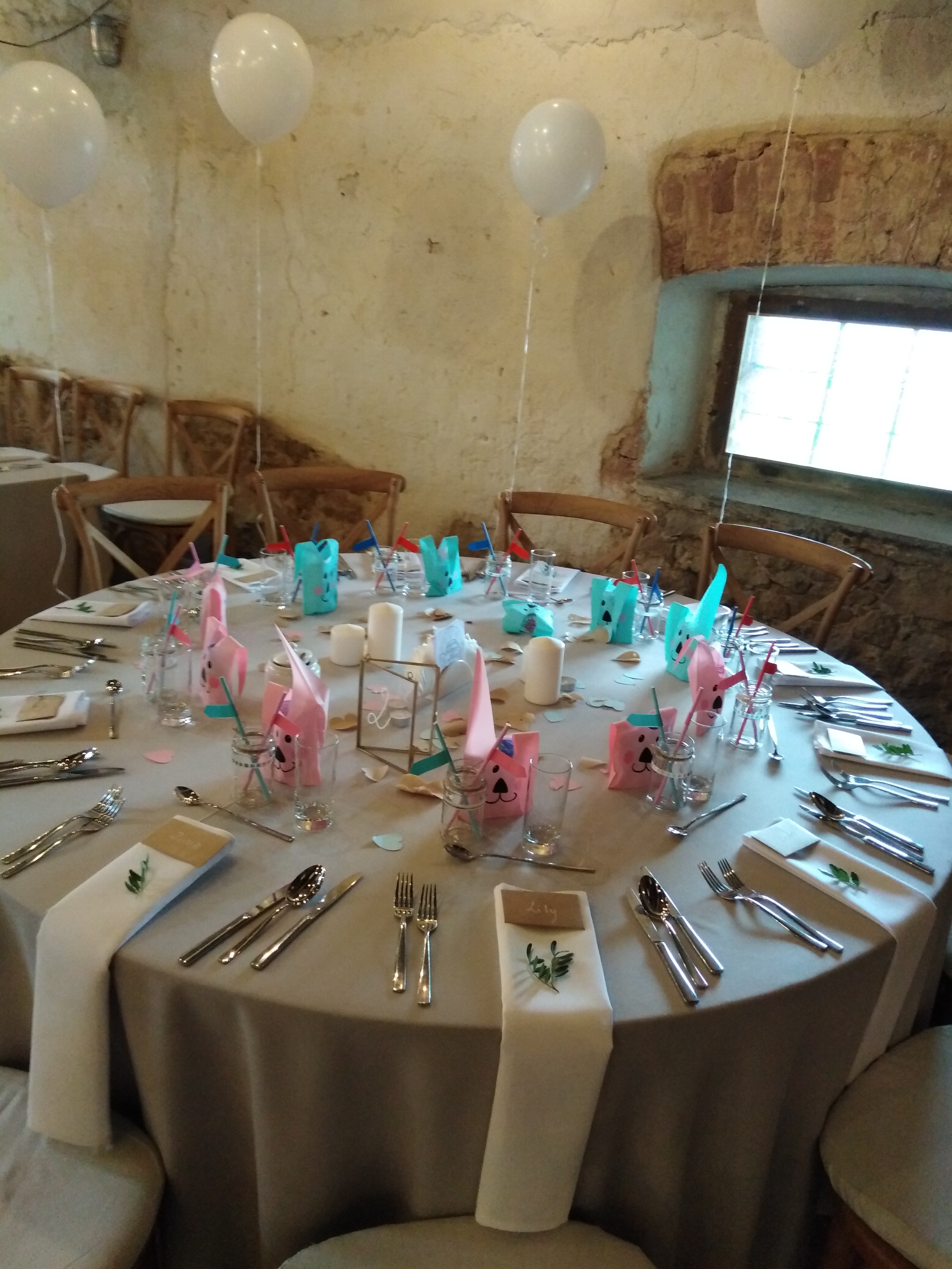 Okrągły stół z szarym obrusem, nakryty dla gości z białymi serwetkami ozdobionymi gałązkami, szklankami, sztućcami, świecami i dekoracjami w kształcie zwierząt, nad stołem unoszą się białe balony.