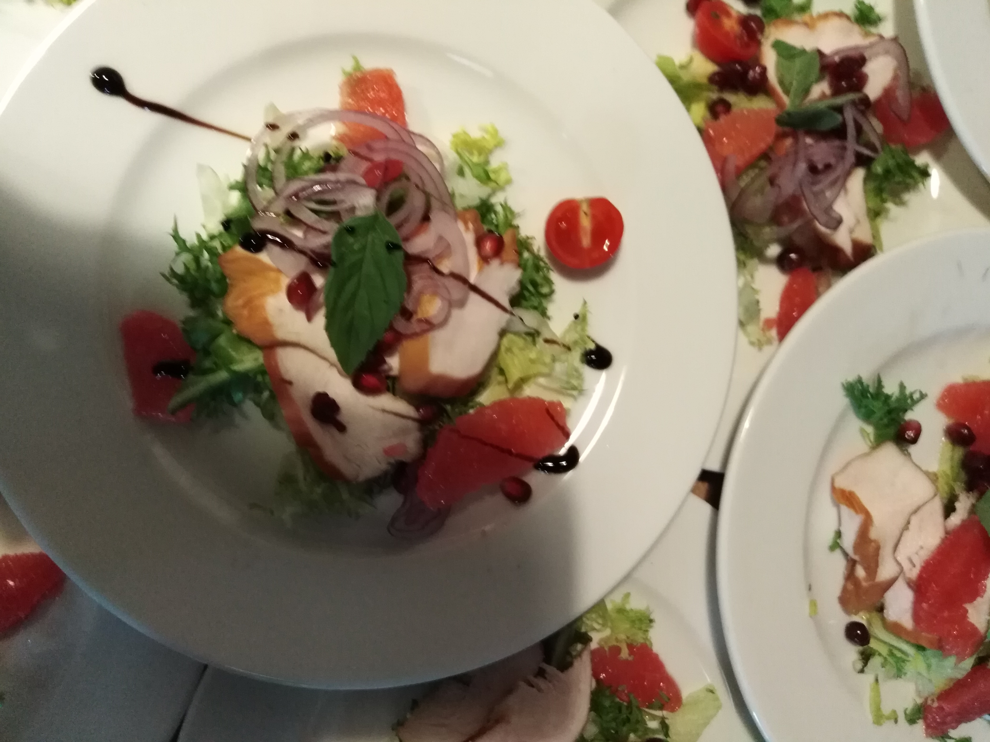 Elegancko ułożona sałatka z grillowanym kurczakiem, grejpfrutem, granatem, czerwoną cebulą i bazylią, polana sosem balsamicznym, na białym talerzu. Kilka talerzy z podobną kompozycją w tle.