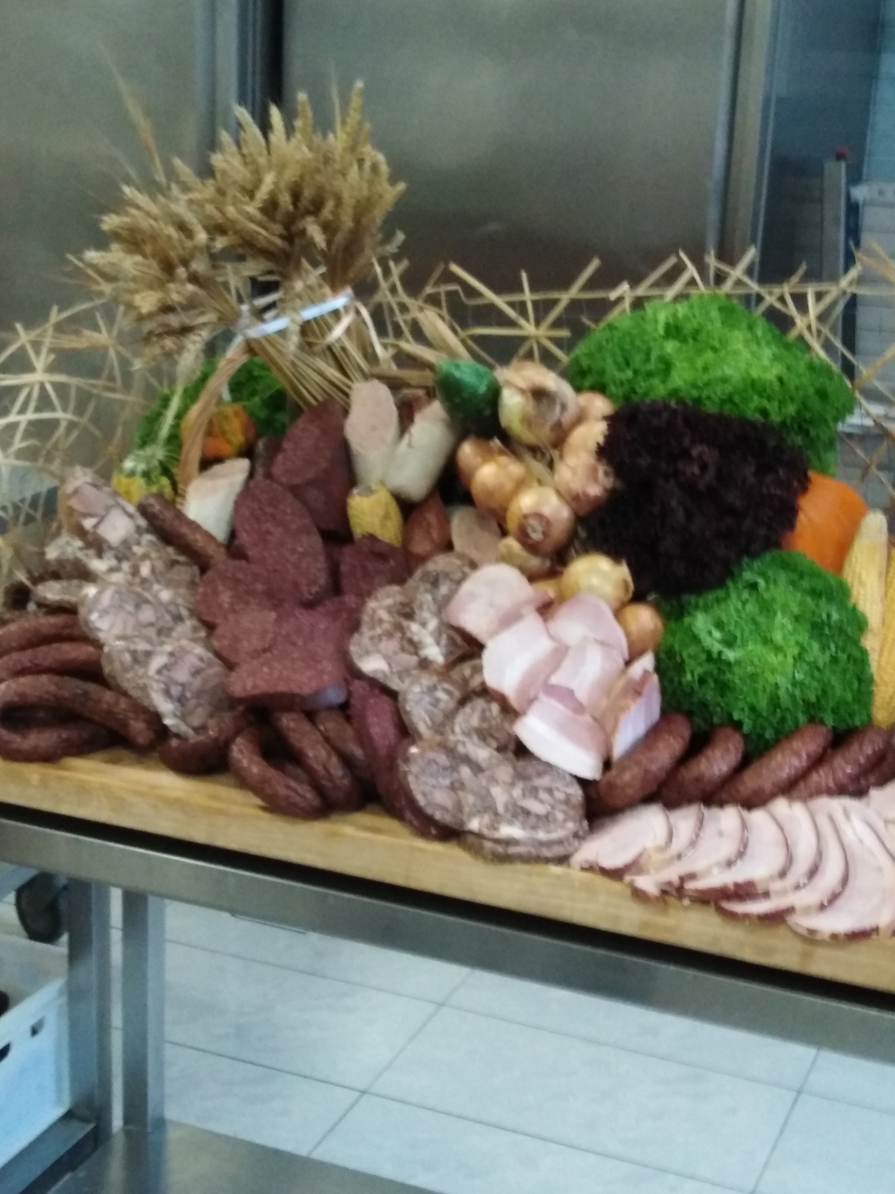 Bogaty stół z wędlinami, kiełbasami, boczkiem, cebulą, sałatą i zbożem, ustawiony na metalowym stole cateringowym.