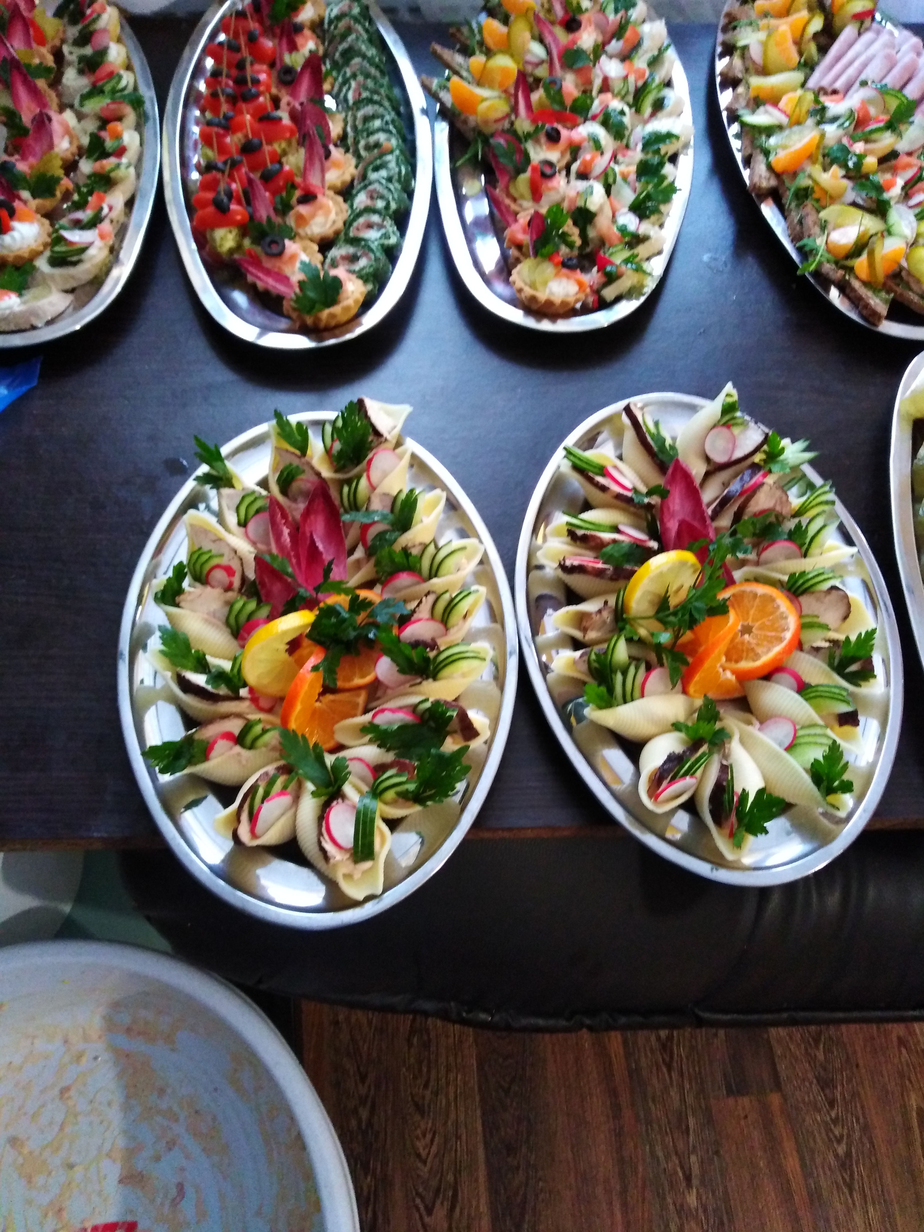 Srebrne tace z dekoracyjnymi przekąskami: makaronowe muszle z farszem, ozdobione plasterkami rzodkiewki, ogórka, pomarańczy i natką pietruszki, oraz tartaletki z pastami i warzywami. Widoczny...