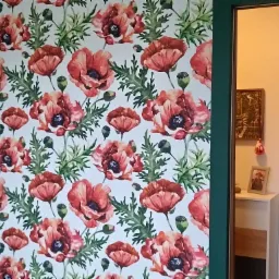 Ściana pokryta tapetą z motywem czerwonych maków i zielonych liści, widoczna z perspektywy wnętrza mieszkania, z fragmentem zielonej framugi drzwi po prawej stronie kadru.
