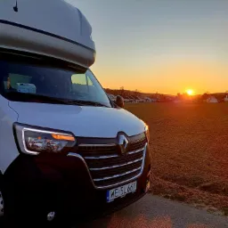 Biały bus Renault z dużą przestrzenią ładunkową na dachu, zaparkowany na tle pomarańczowego zachodu słońca nad polami i zabudowaniami.