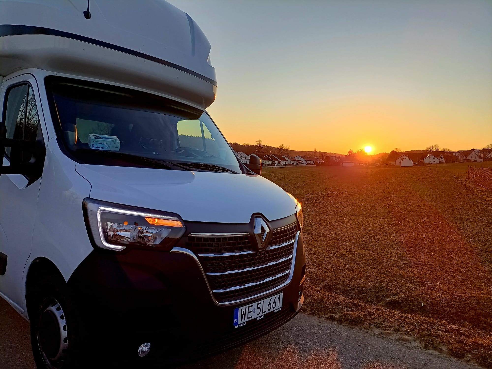 Biały bus Renault z dużą przestrzenią ładunkową na dachu, zaparkowany na tle pomarańczowego zachodu słońca nad polami i zabudowaniami.