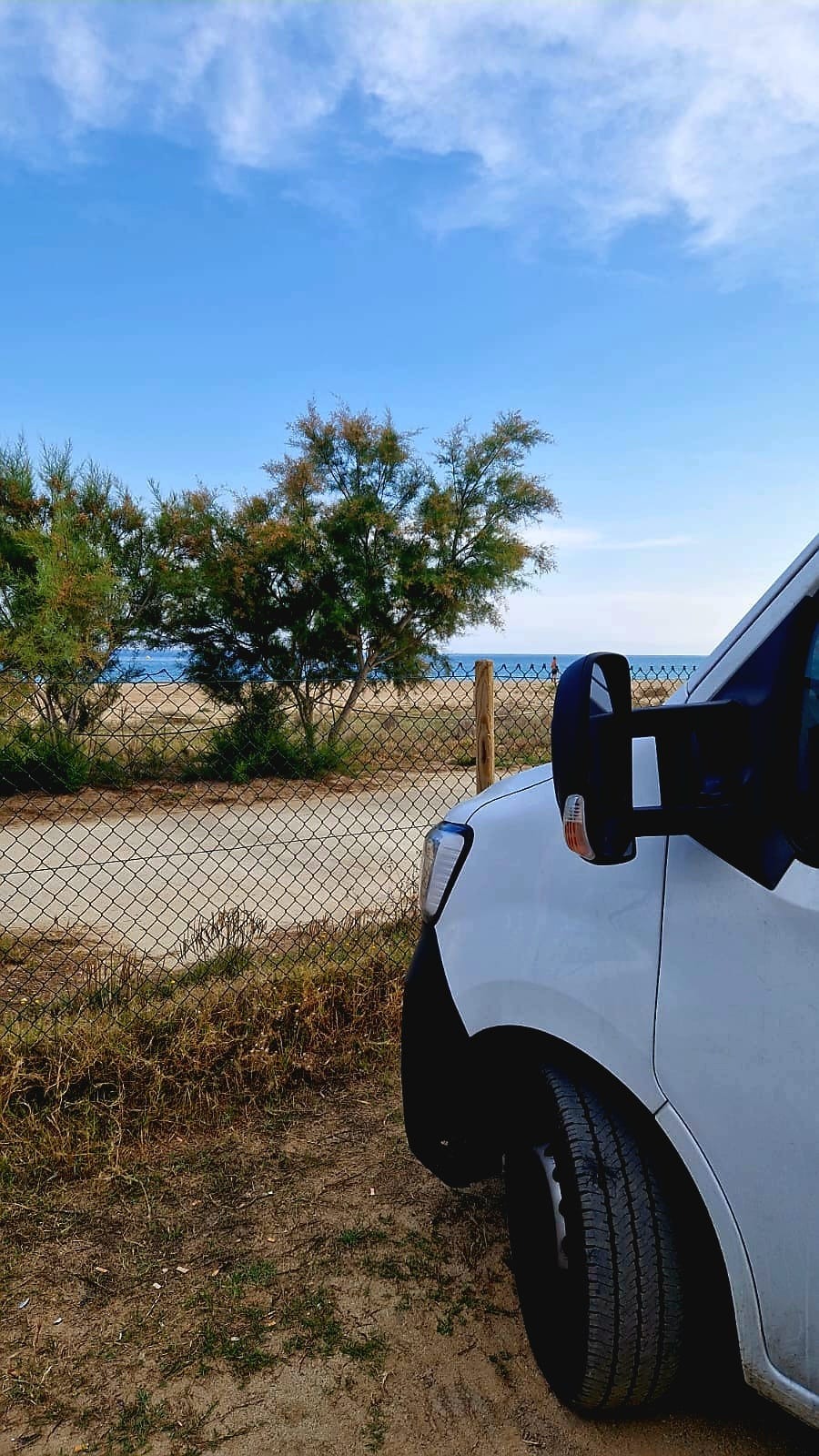 Biały bus transportowy zaparkowany w Bielawie przy piaszczystej drodze w pobliżu plaży i morza, widoczne drzewa i błękitne niebo.