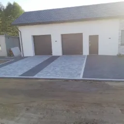 Ułożona kostka brukowa przed garażem dwustanowiskowym z ciemnymi bramami segmentowymi, widoczne pasy z ciemnej kostki oddzielające podjazdy, obrzeża z jasnej kostki, w tle budynek gospodarczy...