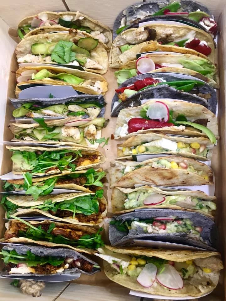 Różnorodne tacos w drewnianej skrzynce, część z jasnymi tortillami, część z ciemnymi, z różnymi farszami i dodatkami: ogórki, rzodkiewki, kukurydza, zielenina, mięso.