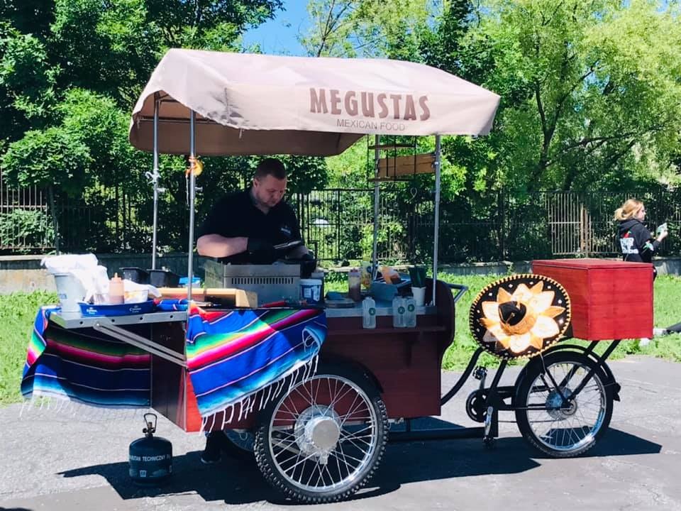 Mobilny wózek z grillem i szyldem 'MEGUSTAS Mexican Food', przygotowujący dania meksykańskie na zewnątrz; kolorowa serweta z frędzlami i dekoracyjny kapelusz sombrero.