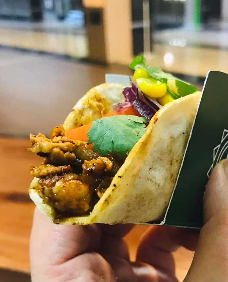 Taco z kurczakiem, kukurydzą, czerwoną kapustą i kolendrą, trzymane w papierowym opakowaniu z logo.