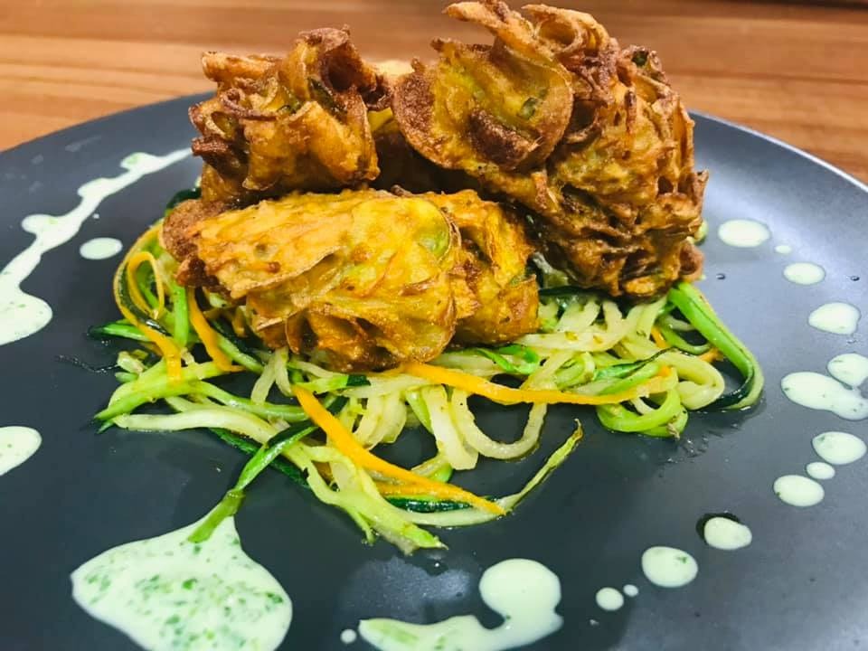 Dekoracyjnie podane smażone kwiaty cebuli na spirali z cukinii i marchewki, z sosem ziołowym na szarym talerzu.