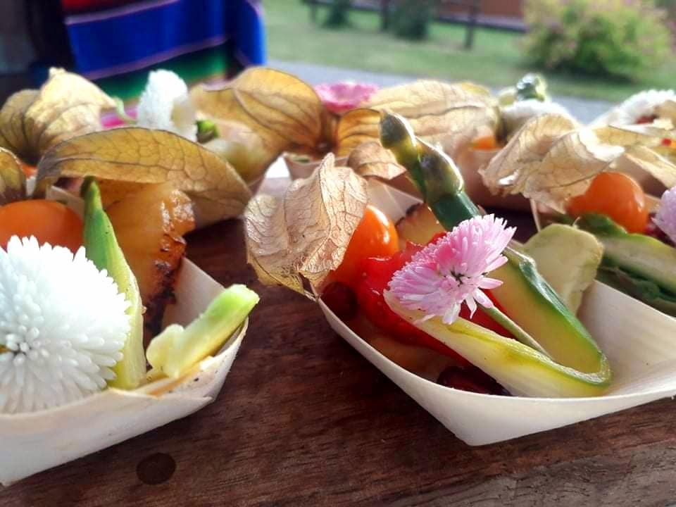 Eleganckie przystawki cateringowe w biodegradowalnych łódeczkach: grillowany ananas, papryka, ogórek i miechunka ozdobione kwiatami, na drewnianym stole.
