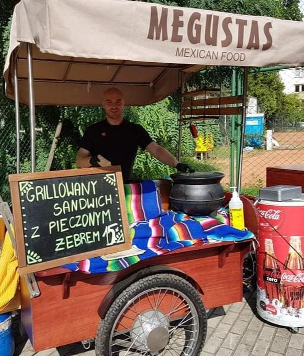 Mobilny wózek gastronomiczny z napisem MEGUSTAS MEXICAN FOOD, oferujący grillowane sandwiche z pieczonym żebrem, obsługiwany przez uśmiechniętego kucharza w czarnych rękawiczkach.
