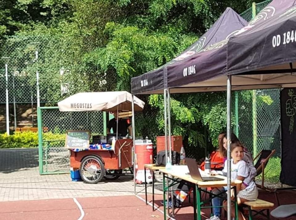 Mobilny wózek cateringowy z napisem MEGUSTAS, zaparkowany obok stanowiska z laptopem i siedzącymi osobami pod namiotem, teren zewnętrzny z zielenią w tle.