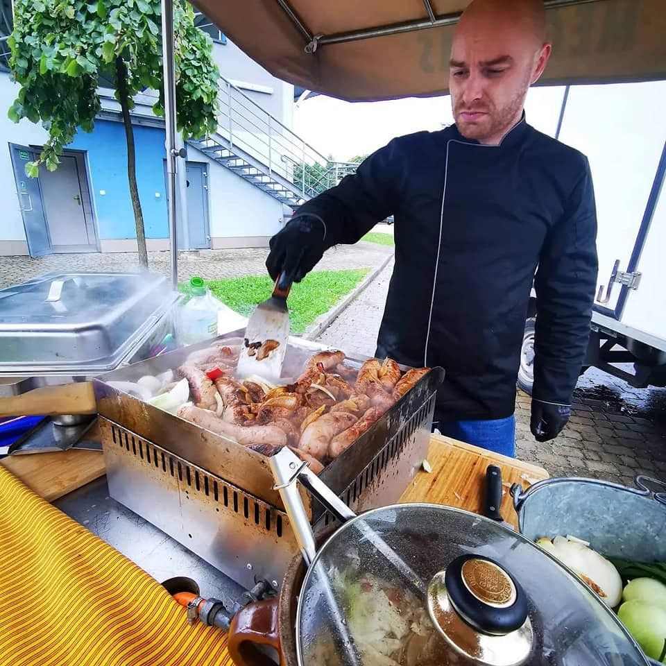 Kucharz w czarnym uniformie, używając łopatki, obraca grillowane mięsa i cebulę na przenośnym grillu podczas imprezy plenerowej.
