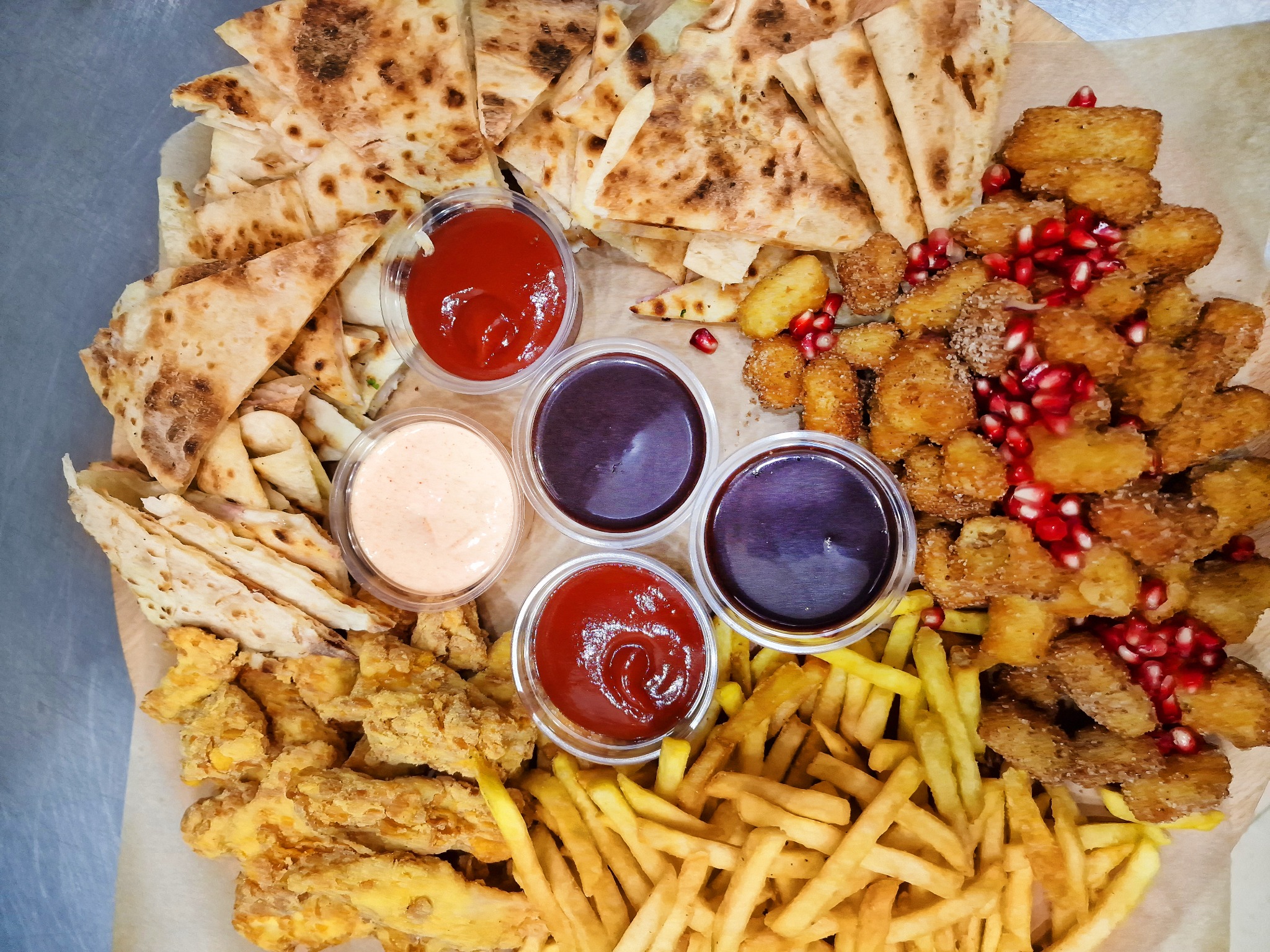 Zestaw przekąsek na drewnianej tacy: trójkąty tortilli, frytki, nuggetsy, churros z posypką z granatu i sosy w plastikowych pojemnikach.