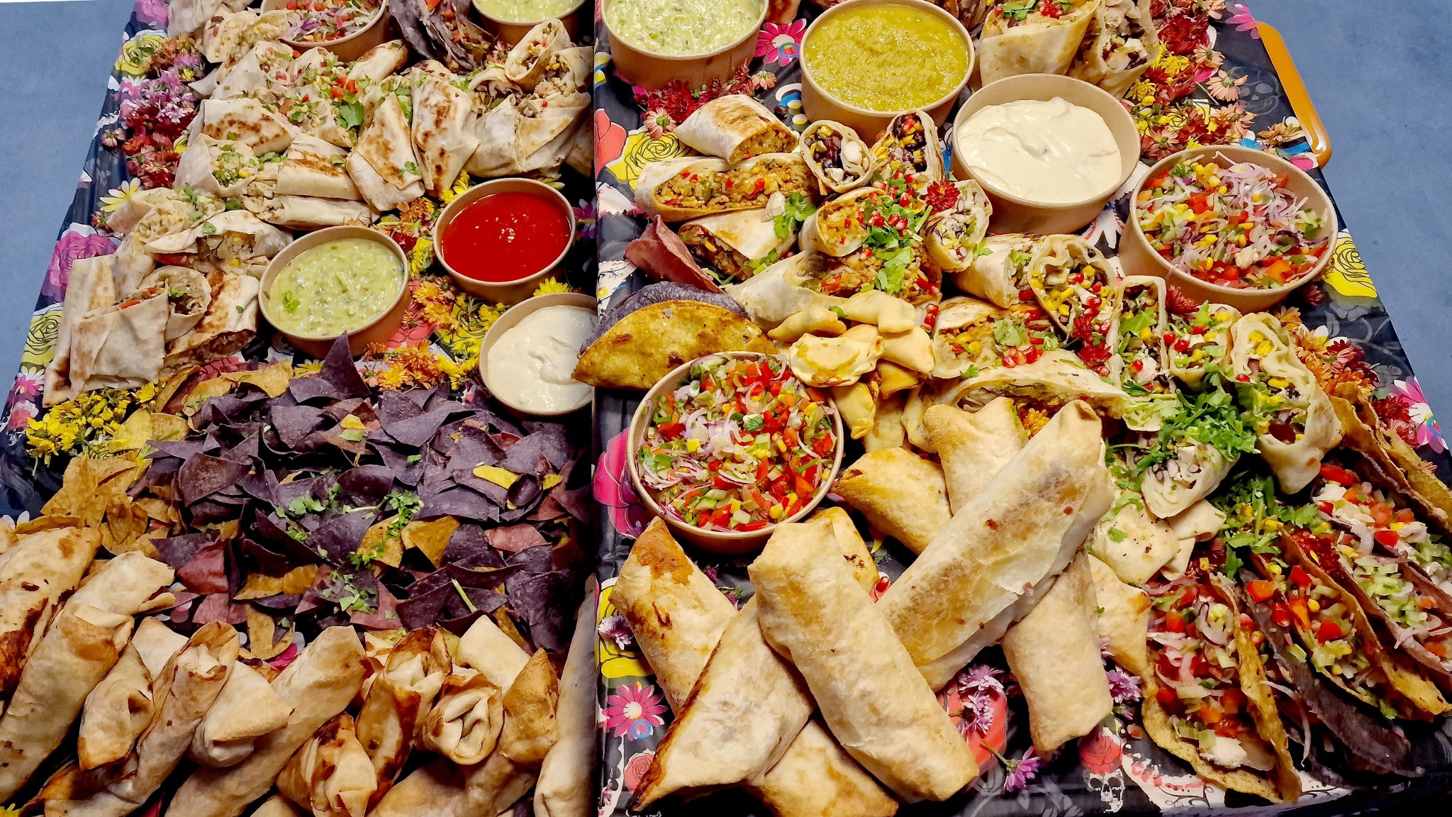 Bogato zastawiony stół z różnorodnymi meksykańskimi przekąskami: burrito, tacos, quesadillas, nachosy z kolorowymi sosami i świeżymi warzywami, ułożonymi na dekoracyjnej tacy z motywem kwiatowym...