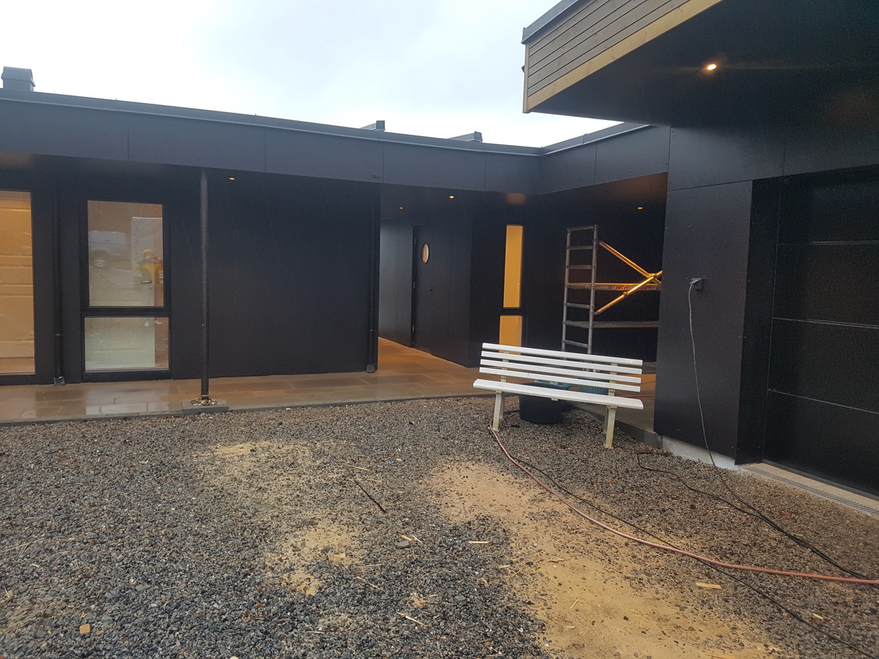 Nowoczesny dom z czarną elewacją, widoczna biała ławka i drabina oparta o ścianę, teren wokół domu wysypany żwirem, pochmurne niebo.