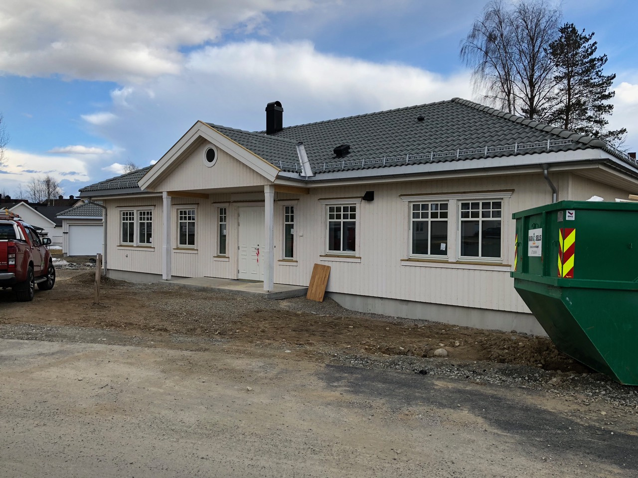 Nowo wybudowany, parterowy dom z białego drewna z szarym dachem, widoczny z zewnątrz, z zielonym kontenerem na śmieci po prawej stronie i czerwonym pickupem w oddali po lewej stronie.