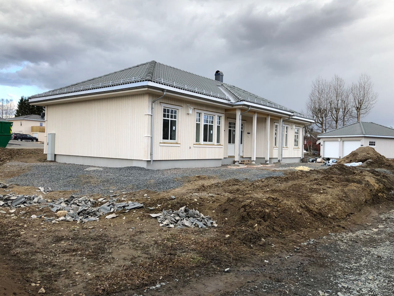 Parterowy dom z jasną elewacją i szarym dachem w trakcie budowy, widoczne przygotowane podłoże i garaż w tle.