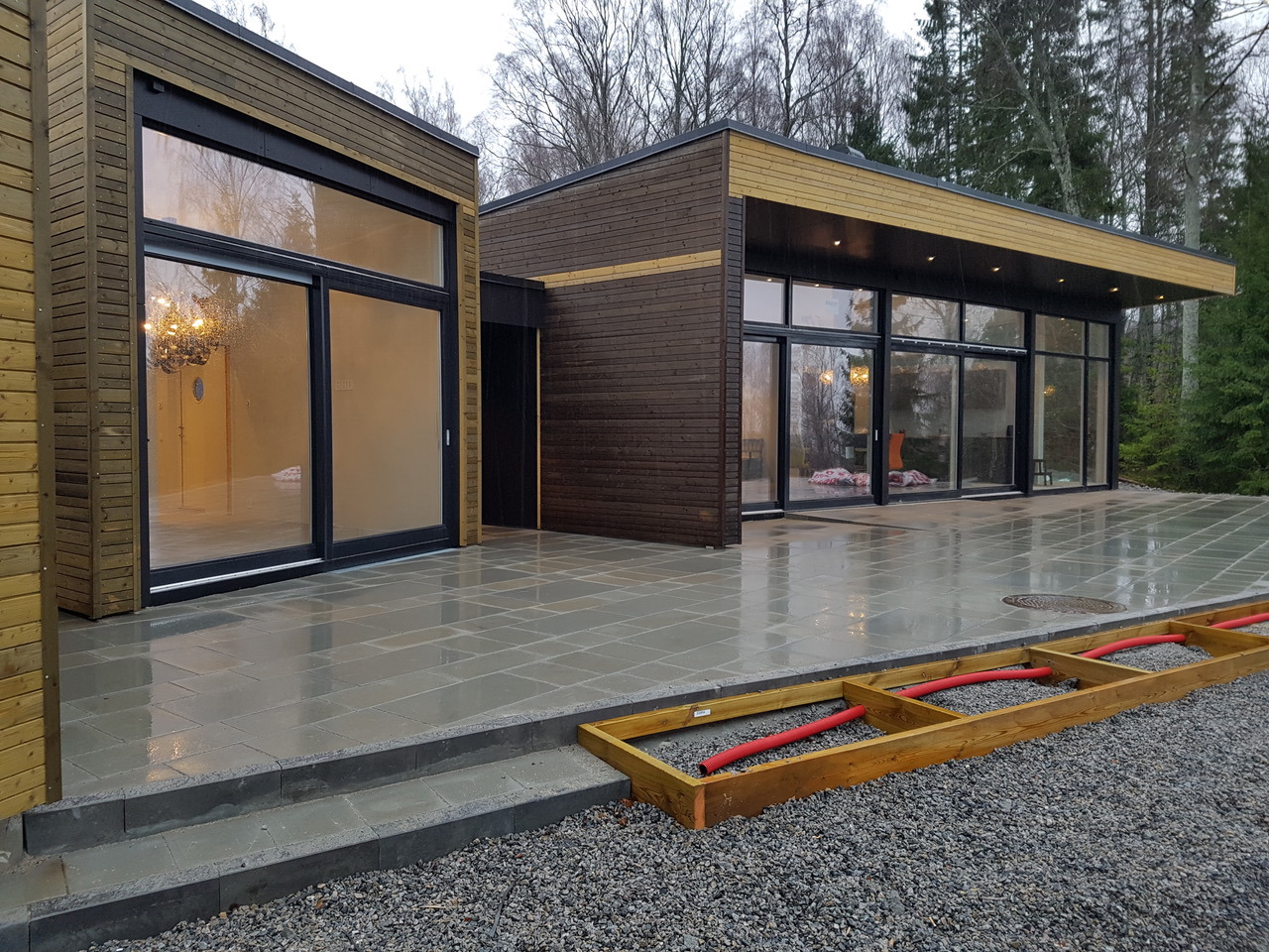 Nowoczesny dom szkieletowy z dużymi przeszkleniami i tarasem wyłożonym szarą kostką, widoczne przygotowania do instalacji ogrzewania podłogowego na zewnątrz.