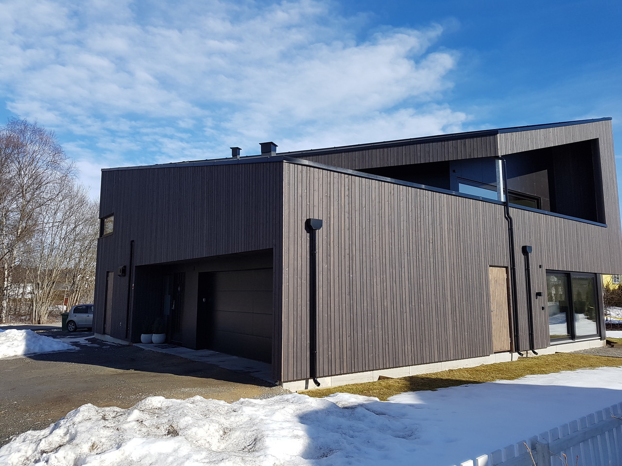 Nowoczesny dom z szarą drewnianą elewacją, z garażem i balkonem, na tle błękitnego nieba i resztek śniegu.