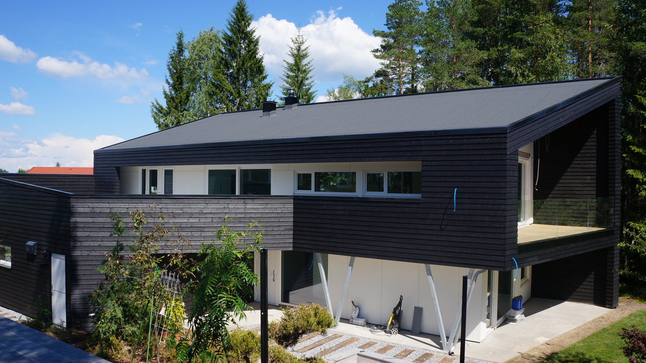 Nowoczesny dom z czarną drewnianą elewacją i płaskim dachem, wsparty na białych, skośnych podporach, z widocznym fragmentem tarasu z przeszkloną balustradą.