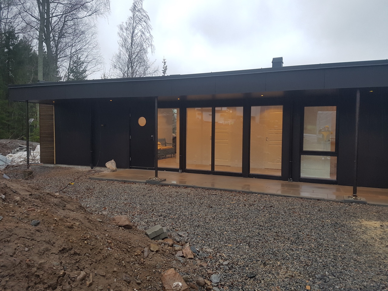 Nowoczesny, parterowy budynek z czarną fasadą i dużymi oknami, widoczny w pochmurny dzień, z niezagospodarowanym terenem wokół.