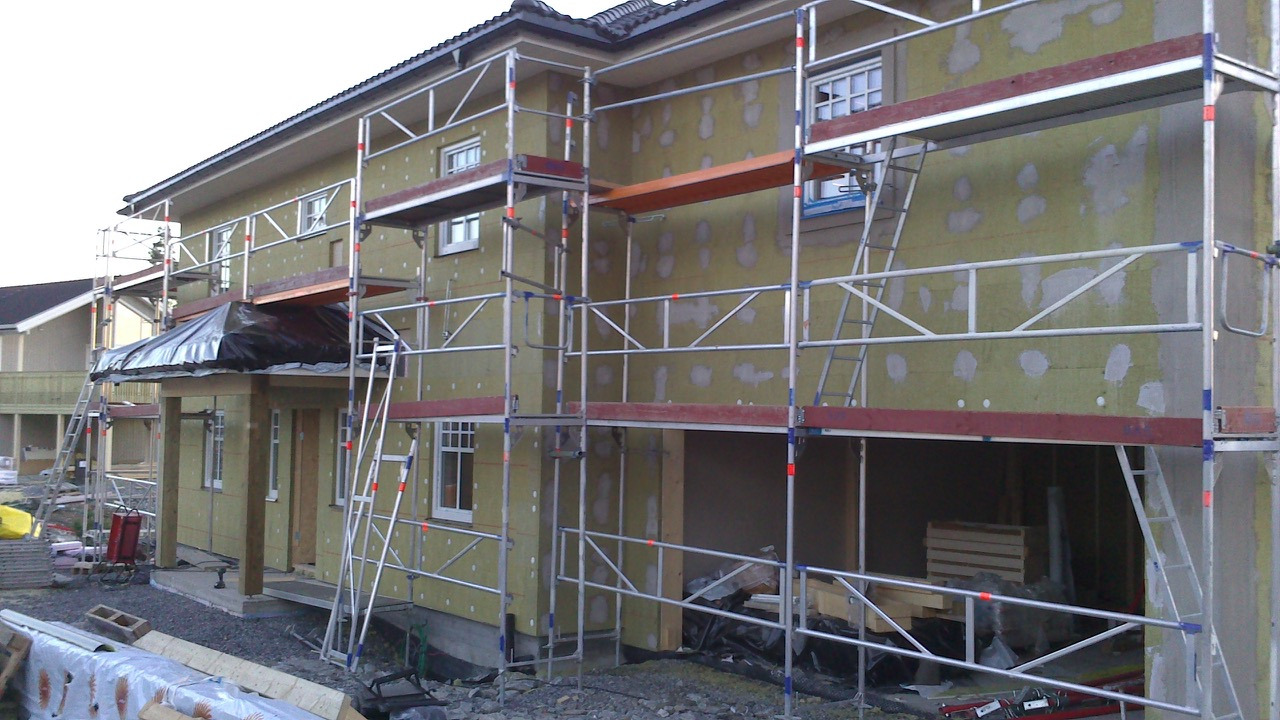 Budynek w trakcie ocieplania, widoczne rusztowania, okna, izolacja ze śladami kleju, częściowo zadaszone wejście.