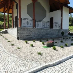 Jasna kostka brukowa i kamienny murek z balustradą otaczają budynek z drewnianą konstrukcją dachu. Ogród z nasadzeniami z przodu. Widok na cmentarz w tle.