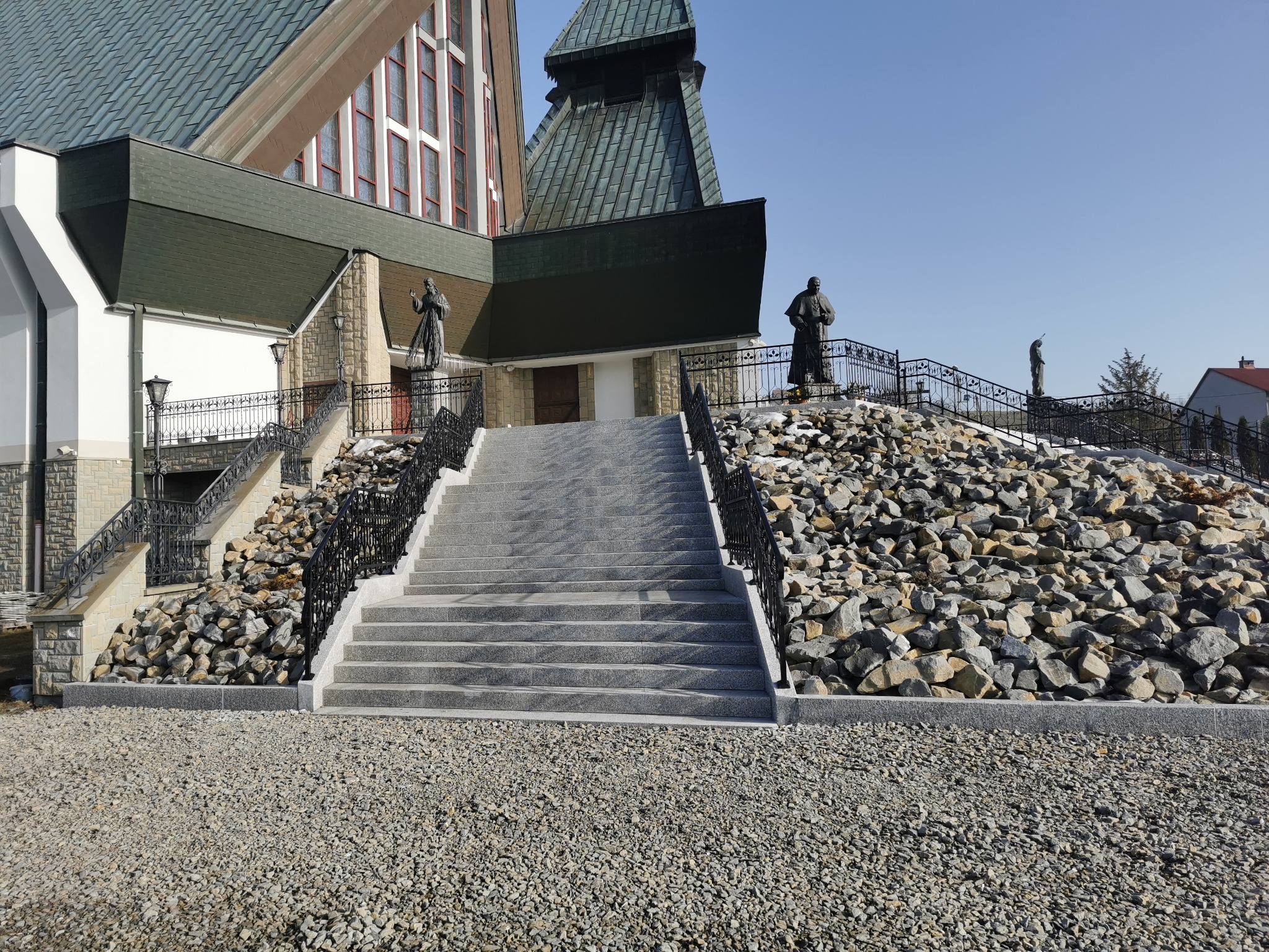 Szerokie, granitowe schody prowadzące do kościoła, otoczone kamieniami i ozdobnymi, kutymi balustradami, z rzeźbami świętych na szczycie.