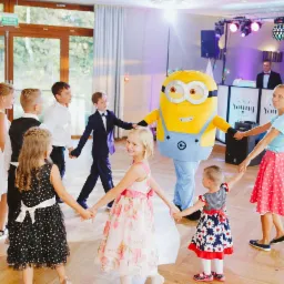 Grupa dzieci trzymających się za ręce w kręgu z osobą w kostiumie Minionka na parkiecie, w tle DJ-ka z oświetleniem na sali imprezowej.