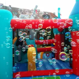 Kolorowy dmuchany zamek otoczony chmurą tęczowych baniek mydlanych, widok na dom w tle.