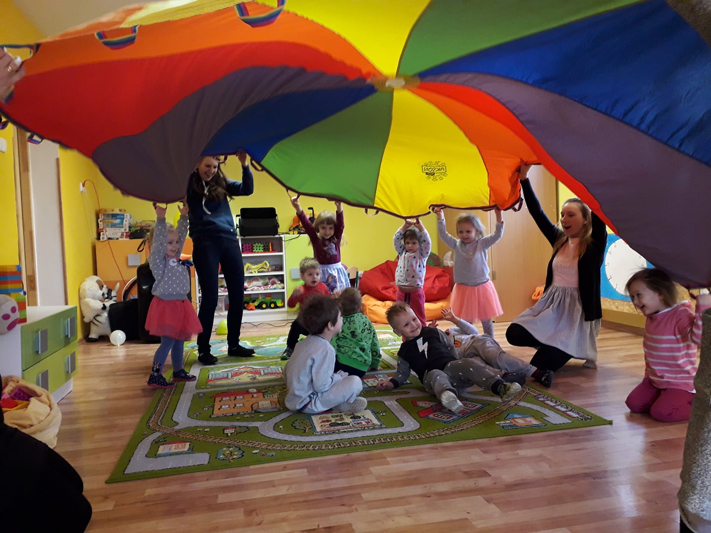 Grupa dzieci i animatorki bawi się kolorową chustą animacyjną w przedszkolu, leżąc i podnosząc ją do góry, widoczny dywan z motywem ulic i budynków.