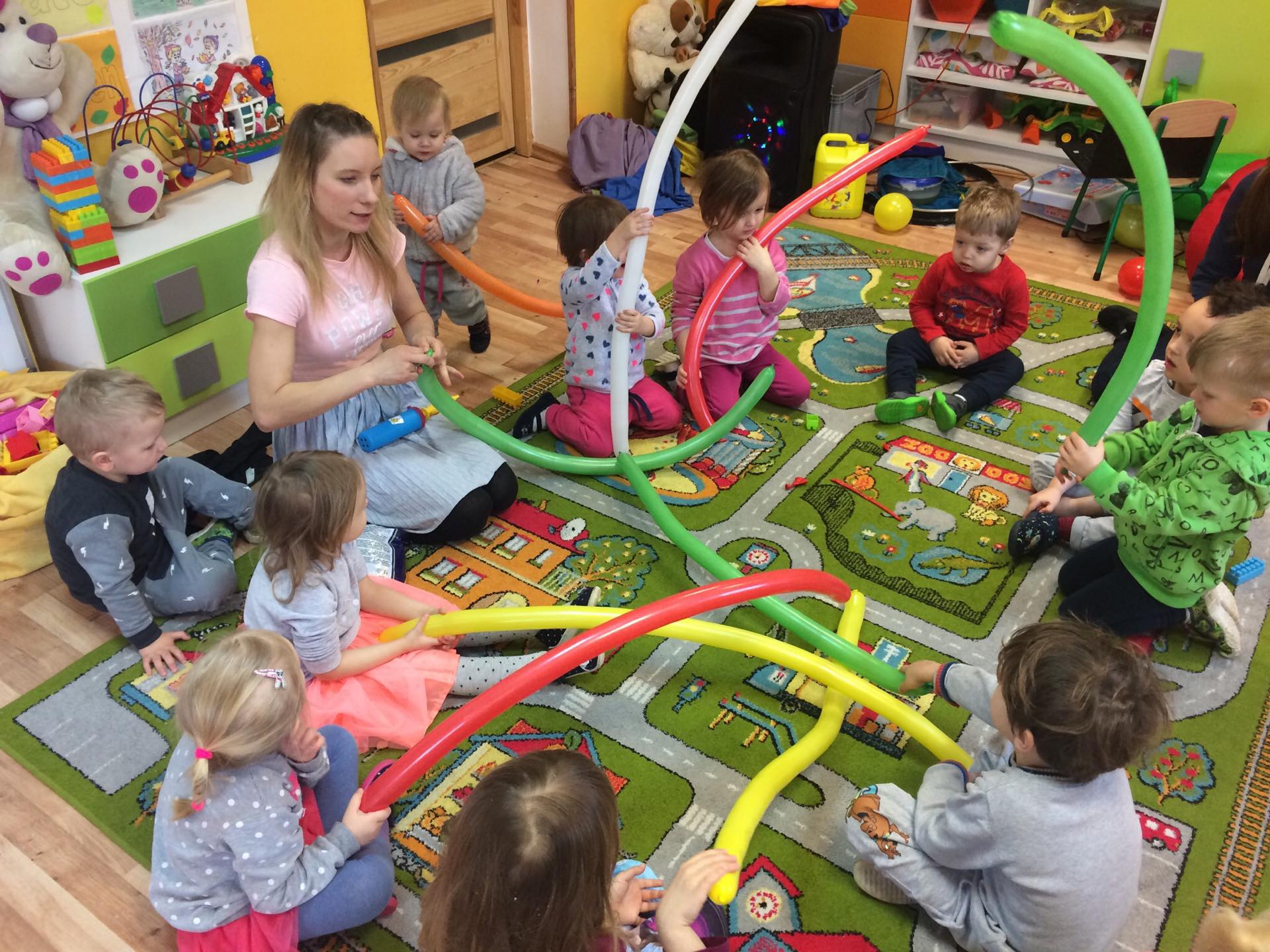 Kobieta w różowej koszulce i szarej spódnicy, siedząca na podłodze z grupą dzieci, pompuje długie balony w różnych kolorach, tworząc z nich kształty na wzorzystym dywanie w pokoju zabaw.