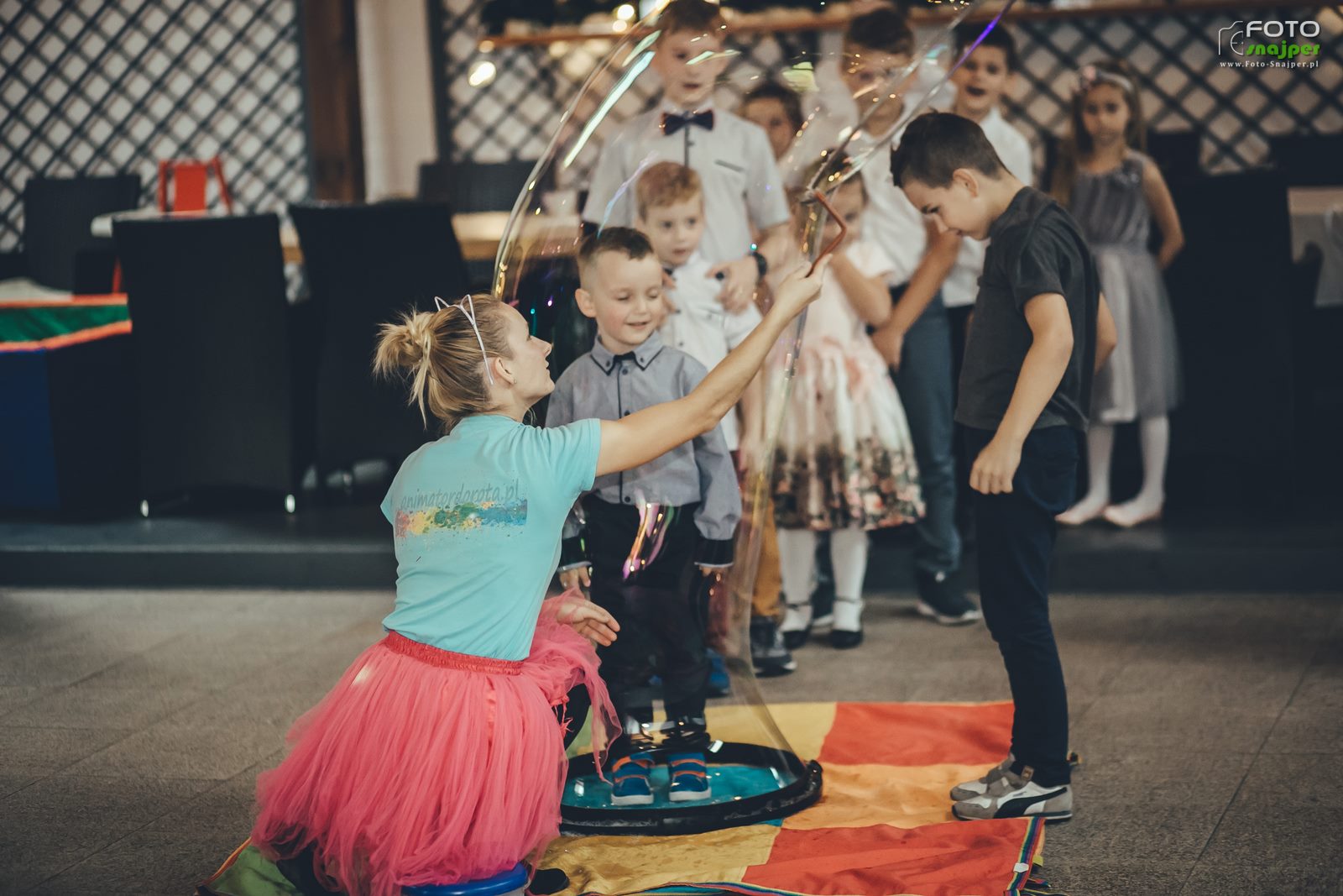 Animator w różowej tiulowej spódnicy robi ogromne bańki mydlane dla grupy dzieci podczas imprezy, na podłodze kolorowa mata.