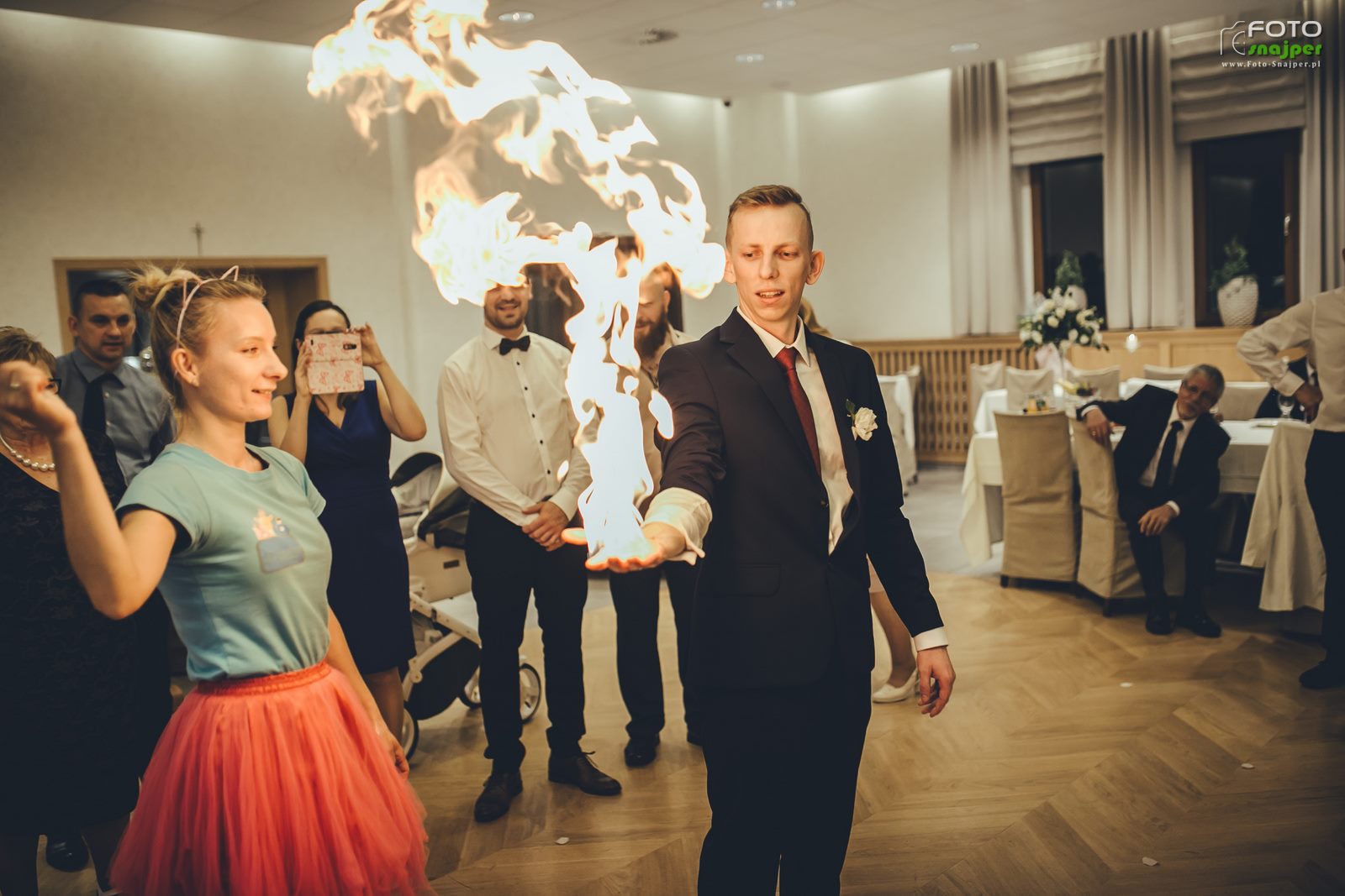 Mężczyzna w garniturze z płomieniem ognia unoszącym się nad dłonią podczas występu na imprezie, w tle goście i wózek dziecięcy.