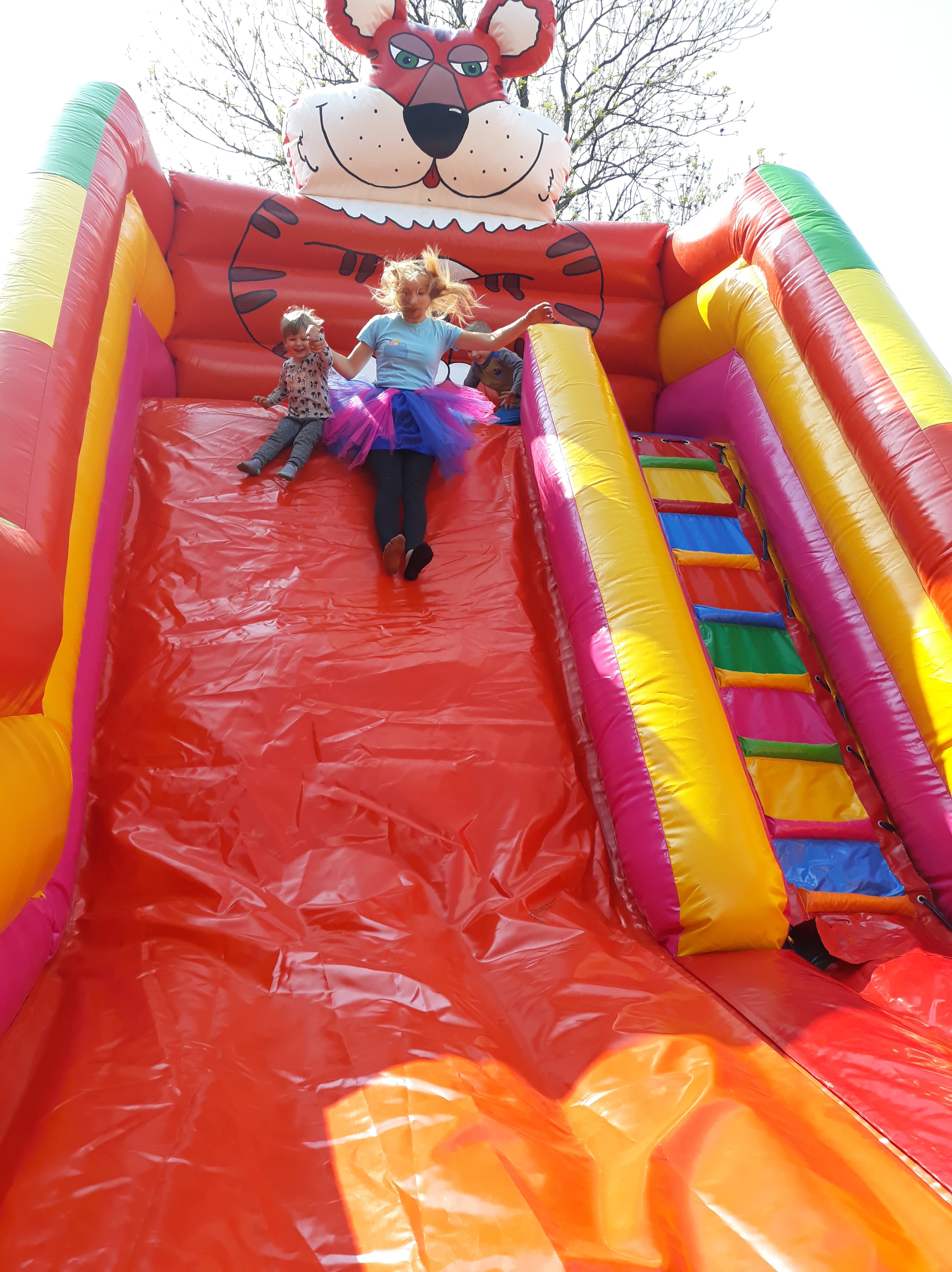 Dzieci z dorosłą osobą zjeżdżają z dmuchanej zjeżdżalni w kształcie tygrysa z kolorowymi elementami.