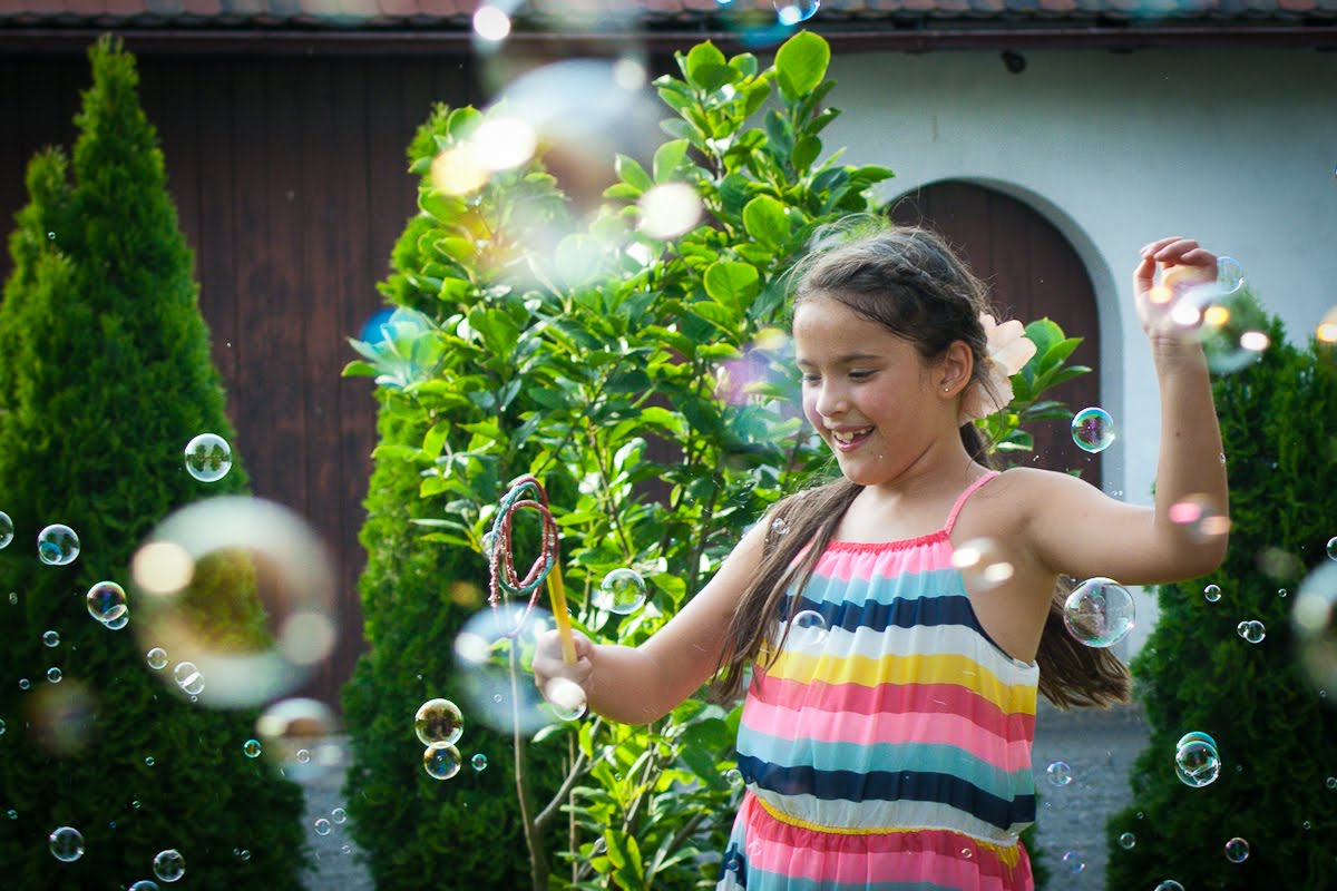 Uśmiechnięta dziewczynka w kolorowej sukience puszcza bańki mydlane w ogrodzie, trzymając w ręku żółtą różdżkę do baniek. W tle zielone krzewy i fragment budynku.