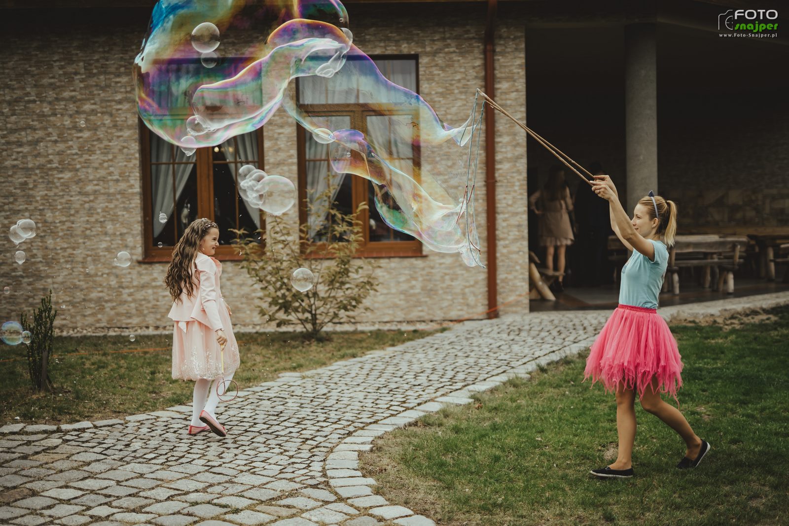 Dziewczynka w różowej sukience obserwuje ogromne bańki mydlane tworzone przez kobietę w różowej spódnicy na brukowanej ścieżce przed budynkiem.