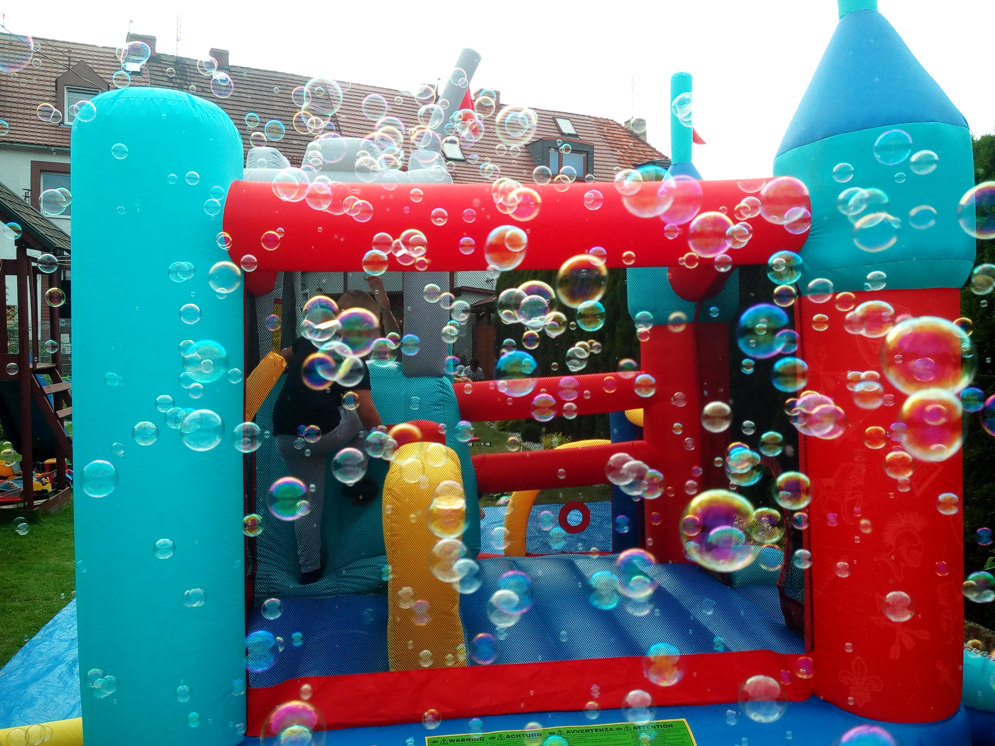 Kolorowy dmuchany zamek otoczony chmurą tęczowych baniek mydlanych, widok na dom w tle.